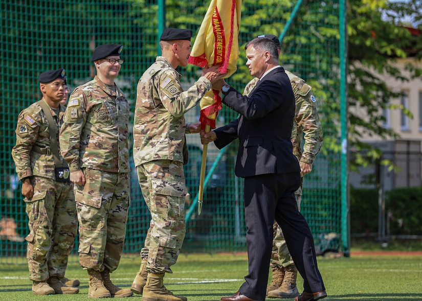 U.S. Army Col. Jeremy McHugh, incoming commander of U.S. Army Garrison Poland, left, takes part in the traditional “passing of the guidon” by receiving the USAG-Poland guidon from Tommy Mize, right, Director of Installation Management Command Europe, during the Garrison’s change of command ceremony, Camp Kościuszko, Poland, July 15, 2025. Established on March 21, 2023, USAG Poland supports 7,500 American Soldiers across 12 sites, delivering quality infrastructure and services to enable V Corps readiness and stands as the Army’s home on the Eastern Flank. (U.S. Army photo by Sgt. Devin Klecan)