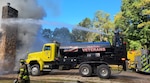 a tanker truck with a yellow cab and a black tank shoots water towards a building on fire.