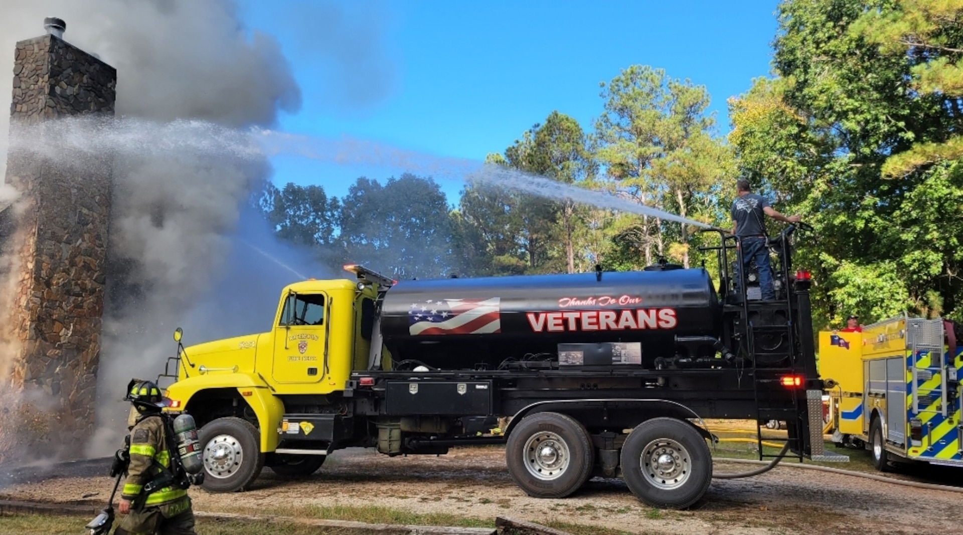 a tanker truck with a yellow cab and a black tank shoots water towards a building on fire.