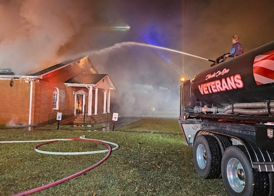 A structure burns in the background of the image while in the foreground the back end of a tanker truck can be seen with a stream of water flying through the air towards the building