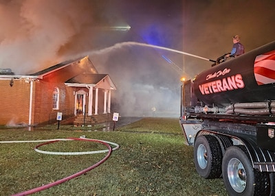 A structure burns in the background of the image while in the foreground the back end of a tanker truck can be seen with a stream of water flying through the air towards the building