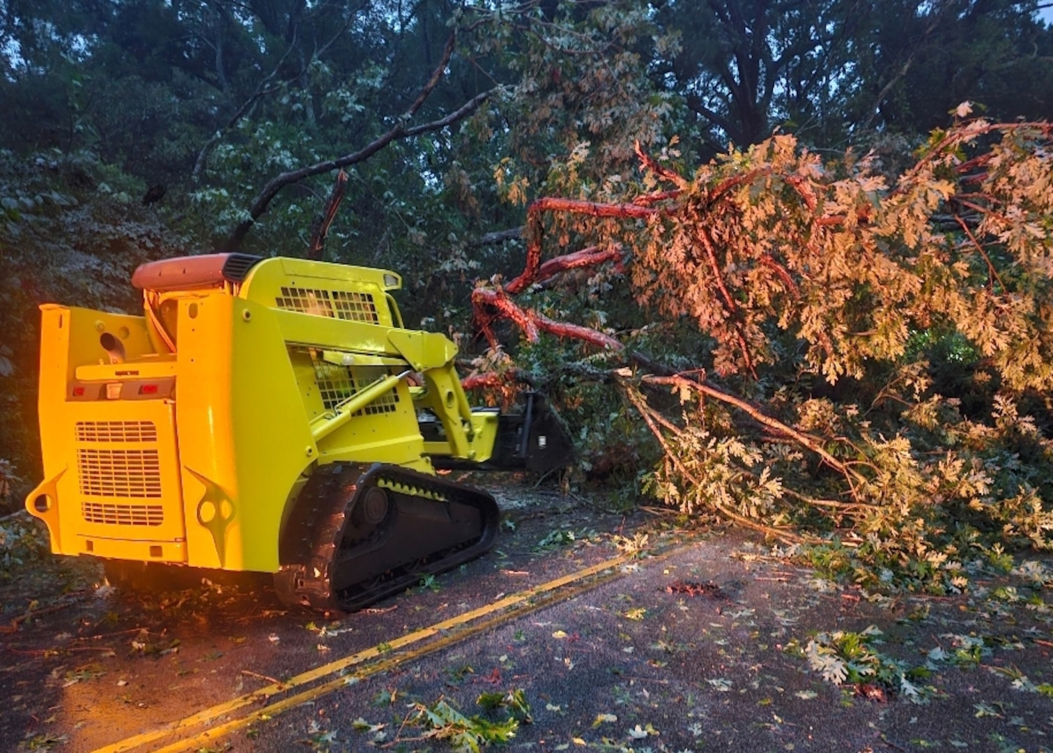 A yellow skid steer faces away from the camera towards a tree that has fallen across the road. it is dark out and the road appears to be wet.