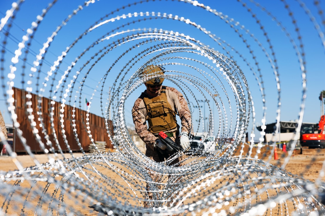 A Marine uses a tool to tie concertina wire together in rolls on a sunny, cloudless day, with a tall security wall and vehicles in the background.