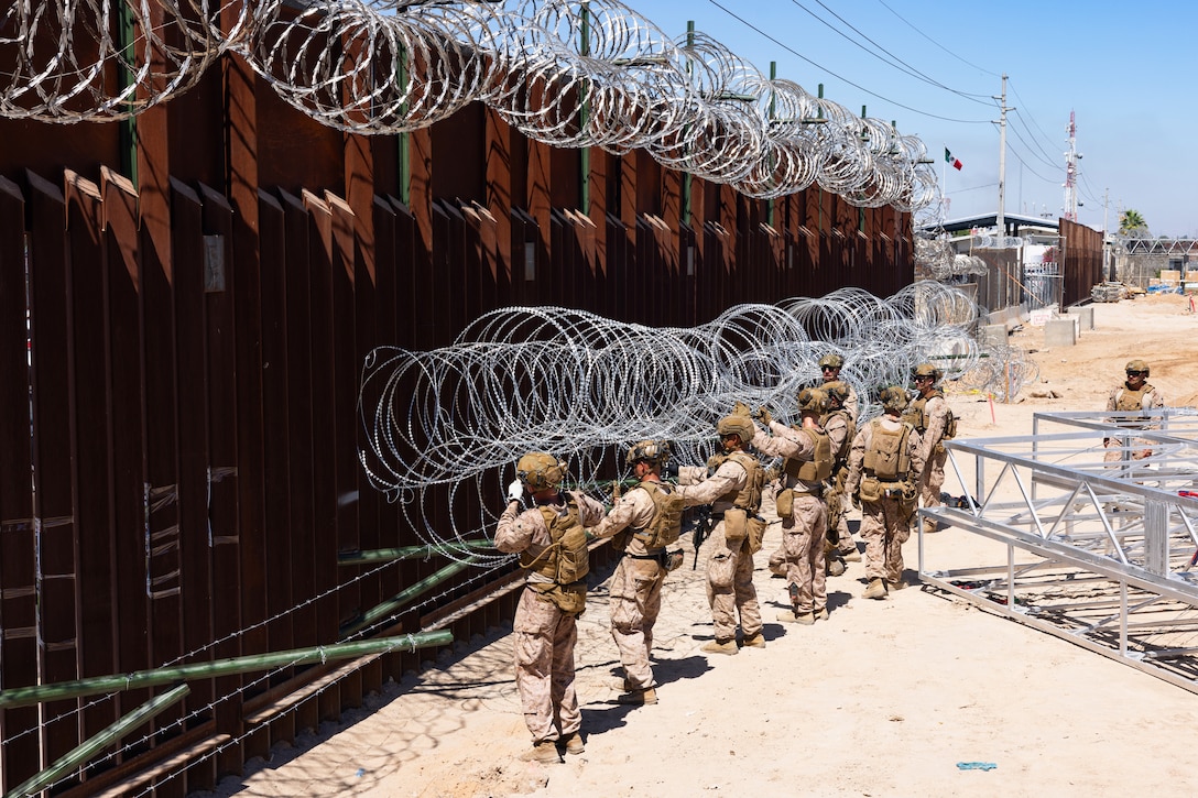 Marines place concertina wire onto rods extending from a tall security barrier in a sandy area on a sunny day, with equipment and a building in the background.