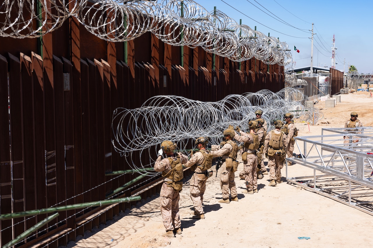 Marines place concertina wire onto rods extending from a tall security barrier in a sandy area on a sunny day, with equipment and a building in the background.