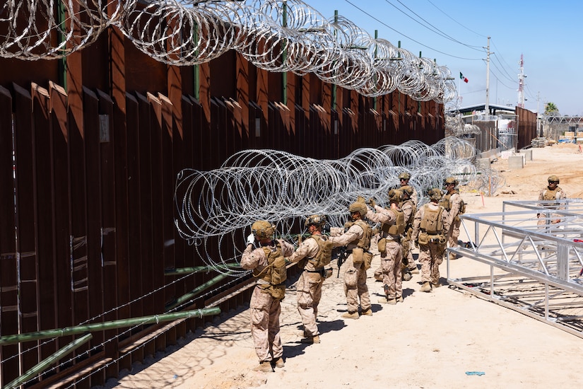 Marines place concertina wire onto rods extending from a tall security barrier in a sandy area on a sunny day, with equipment and a building in the background.