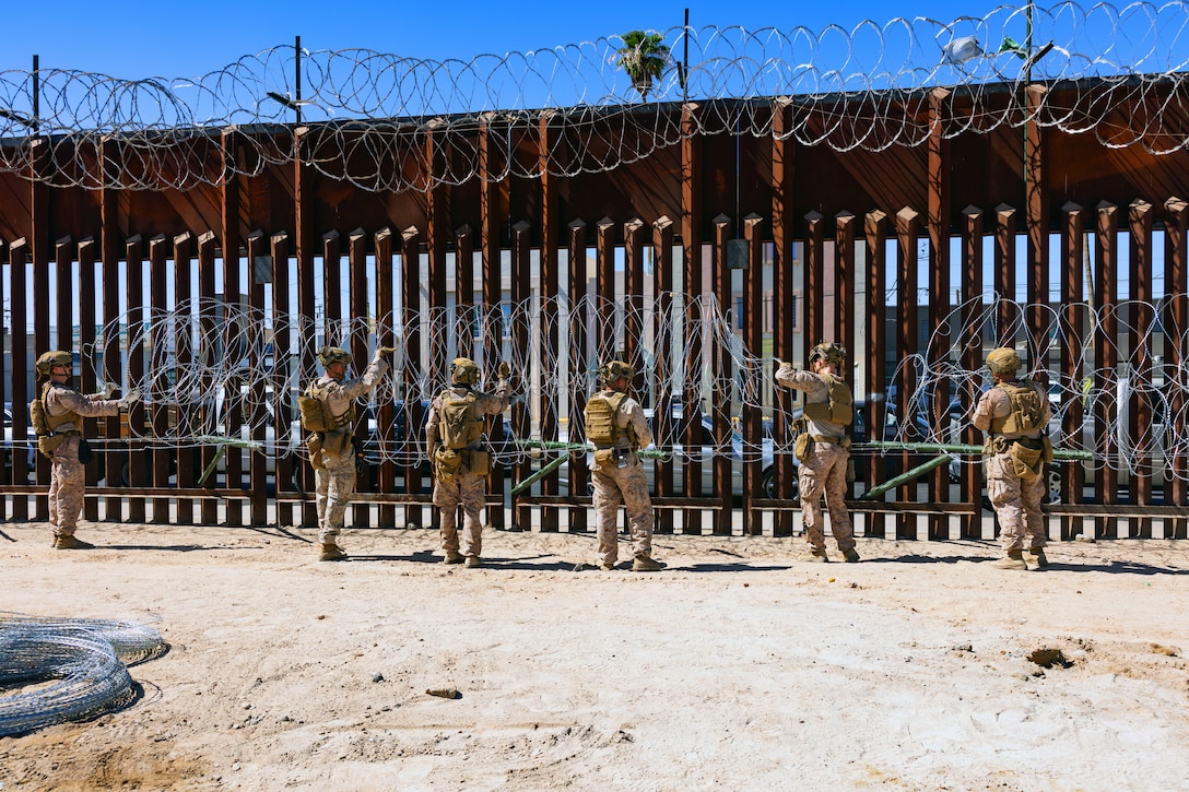 Marines place concertina wire onto rods extending from a tall security barrier in a sandy area on a sunny day.