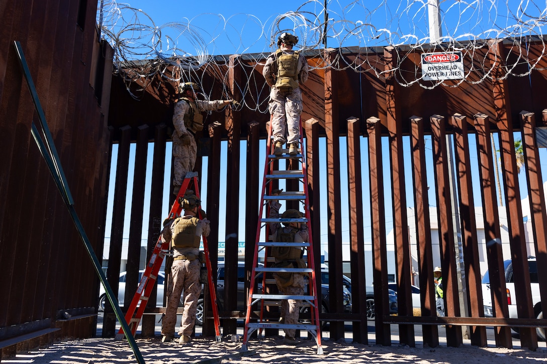 Four Marines, two standing on ladders as two others hold the base of the ladders, work to place concertina wire at the top of a tall, metal barrier on a sunny day.