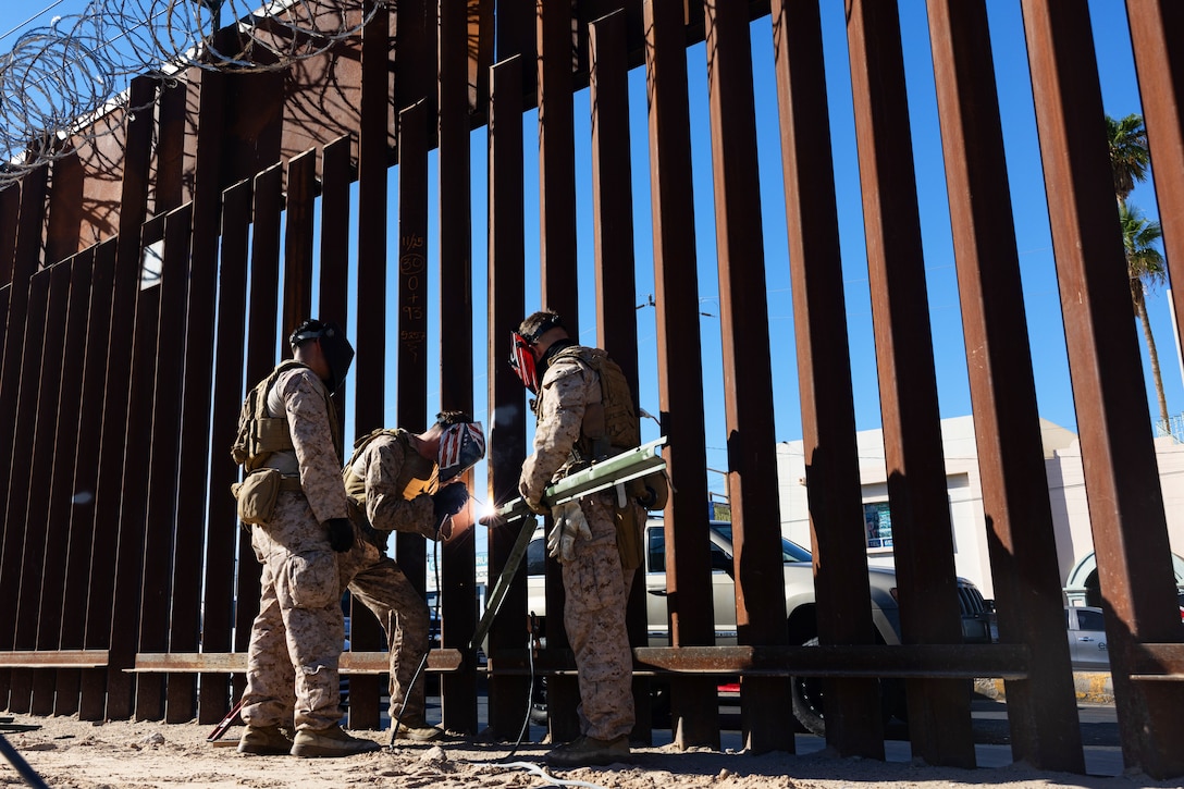 Three Marines, one welding and the other two standing nearby in support, weld brackets to a tall security barrier on a sunny day.
