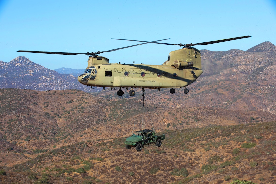 A helicopter carries a green utility vehicle attached by cables to the underside of the helicopter on a sunny day, with mountains in the distance.