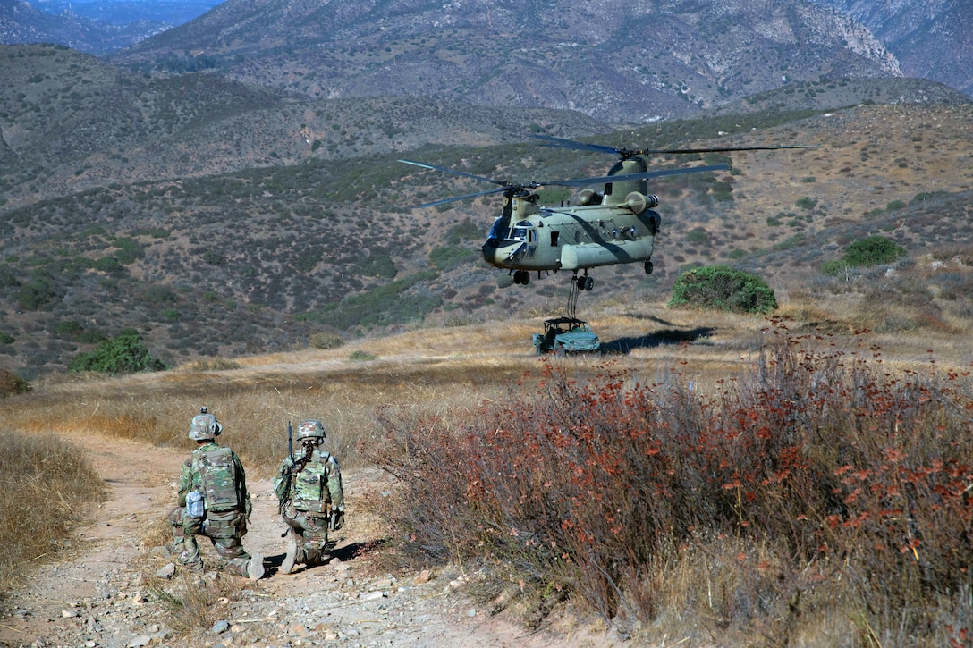 Two soldiers kneel in a sparsely vegetated area as a helicopter hovers nearby, with a vehicle attached by cables on the ground below the helicopter; mountains are in the distance.