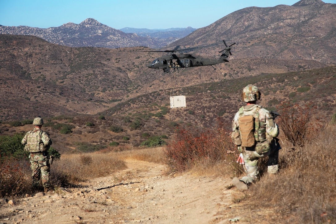 Two soldiers in a sparsely vegetated area watch a hovering Black Hawk deliver a container attached by cables to the helicopter on a sunny day, with mountains in the distance.