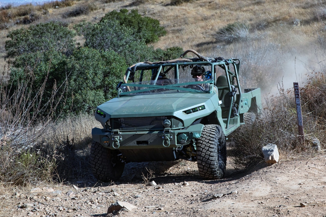 A soldier drives a utility vehicle in a hilly, desert-like area on a sunny day, with sparse vegetation in the background.