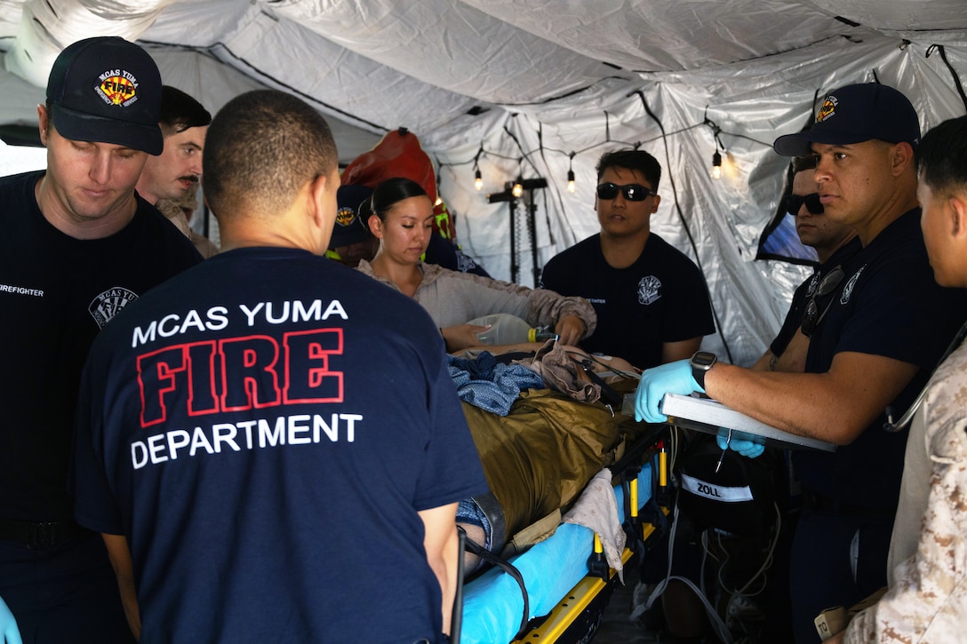 A sailor uses a device to supply air to a simulated casualty victim lying on a stretcher, as other people wearing fire department T-shirts stand and observe in a large tent.