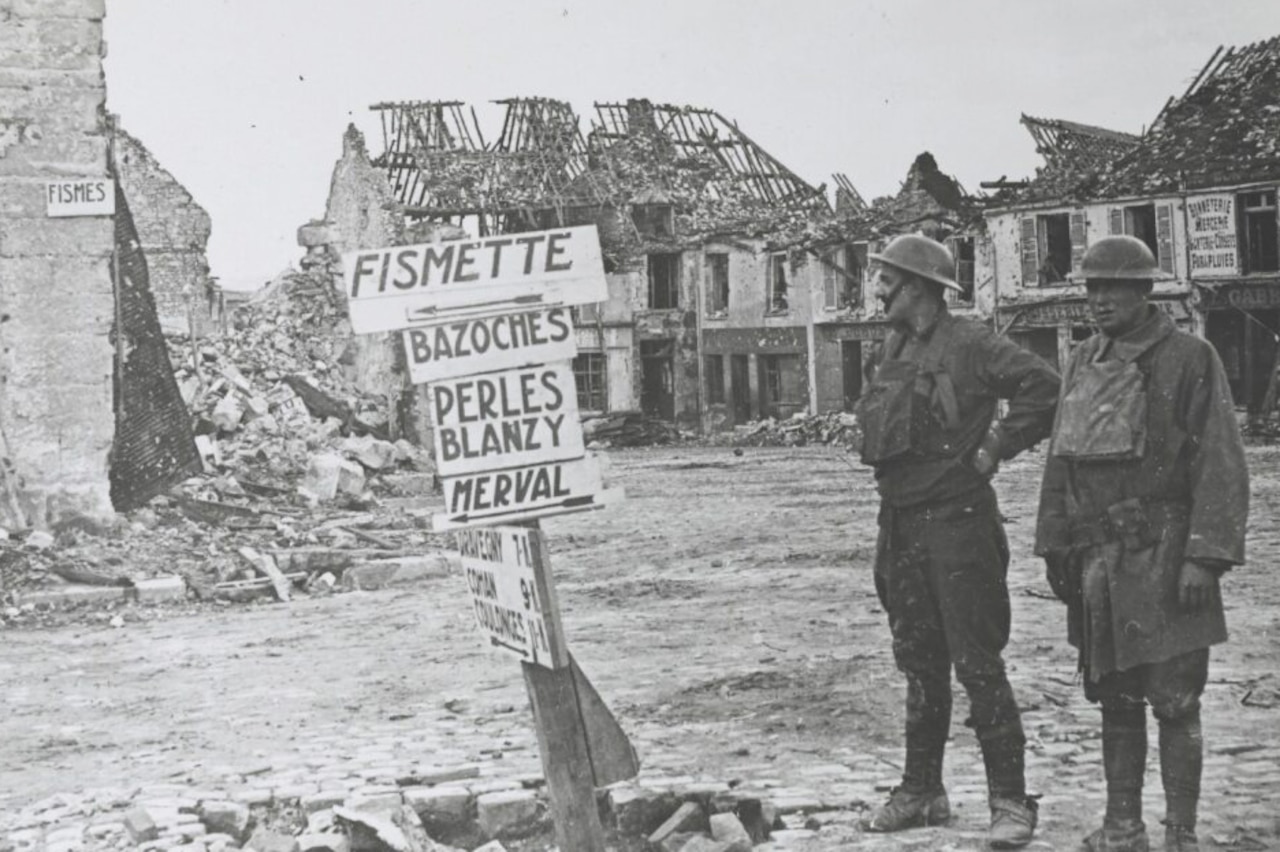 Two soldiers in uniform look at a signpost amid the rubble of a destroyed town.