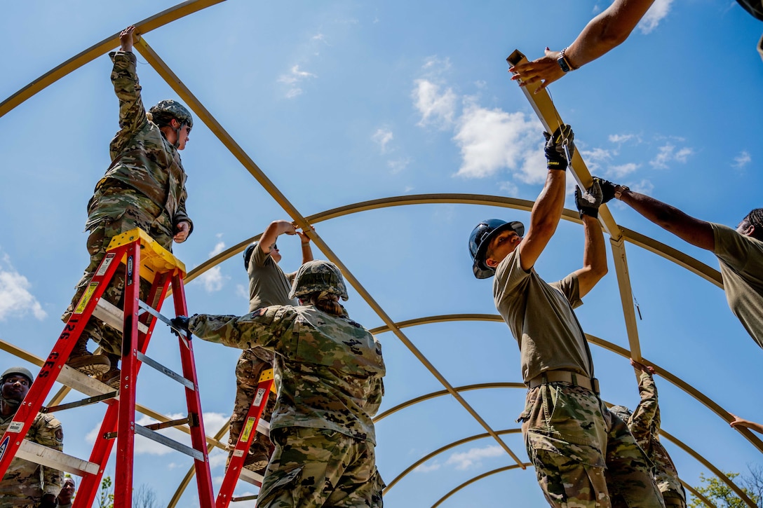 National Guardsmen stand on ladders while building the shell of a tent against a cloudy, blue sky.