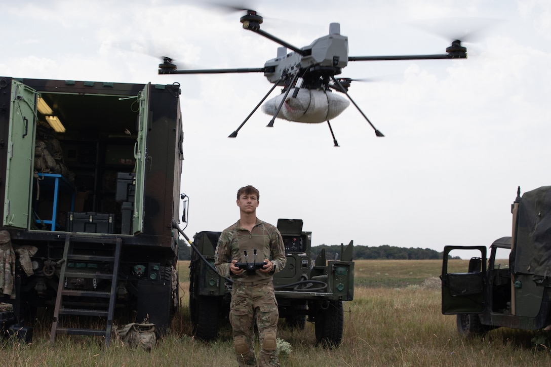 A soldier standing in tall grass in front of two trucks operates an airborne drone.