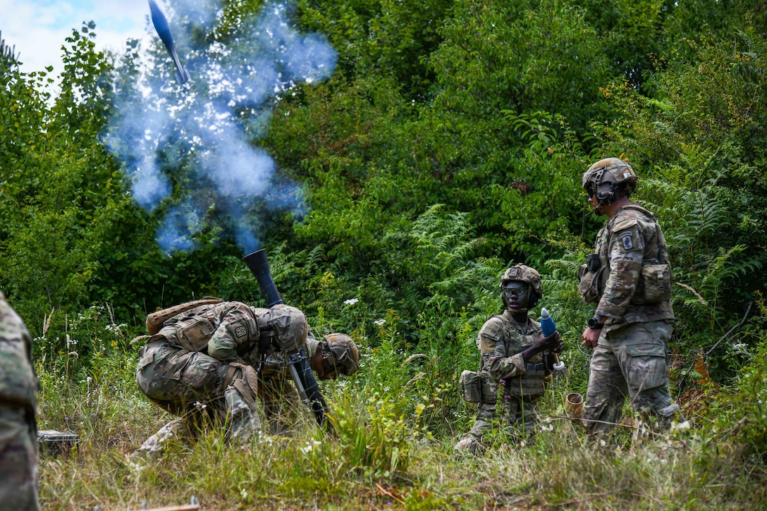 Soldiers in a wooded area fire a weapon creating a cloud of smoke.