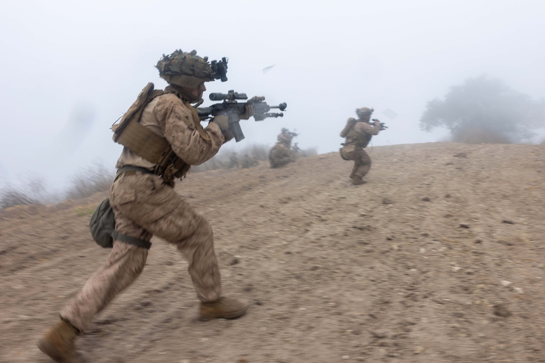 Marines aim weapons while walking up a dirt hill on a foggy day.