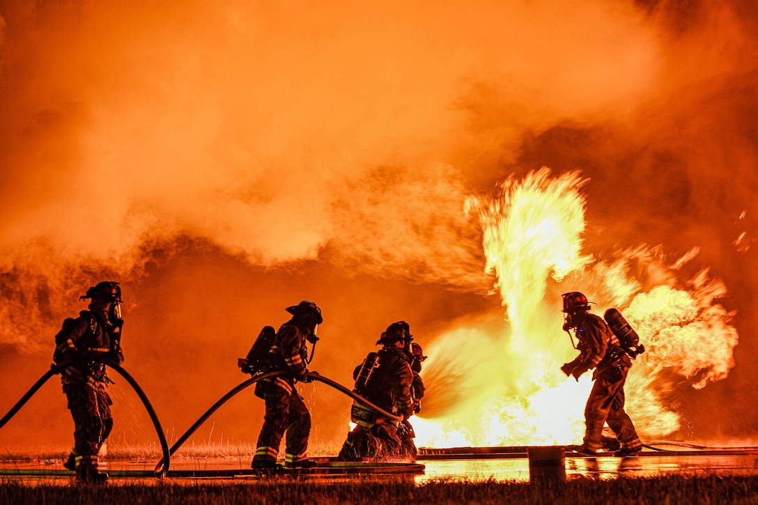 A firefighter watches as four firefighters use a hose to put out a massive fire creating a smoky, orange background.