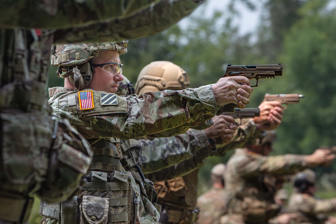 National Guardsmen stand in line and aim weapons against a wooded background.