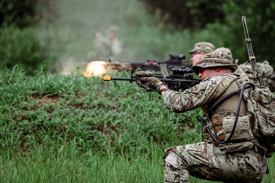 Two kneeling soldiers fire weapons in a wooded area.