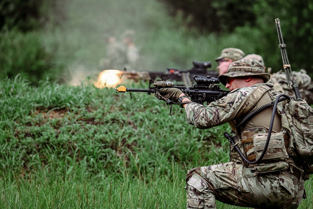 Two kneeling soldiers fire weapons in a wooded area.