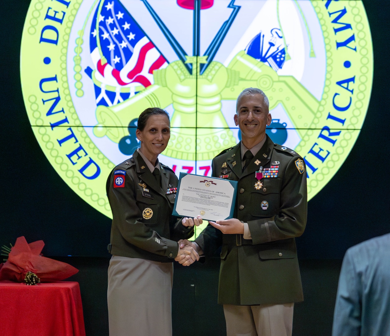 A man and woman in uniform show a certificate while on a stage in an auditorium.