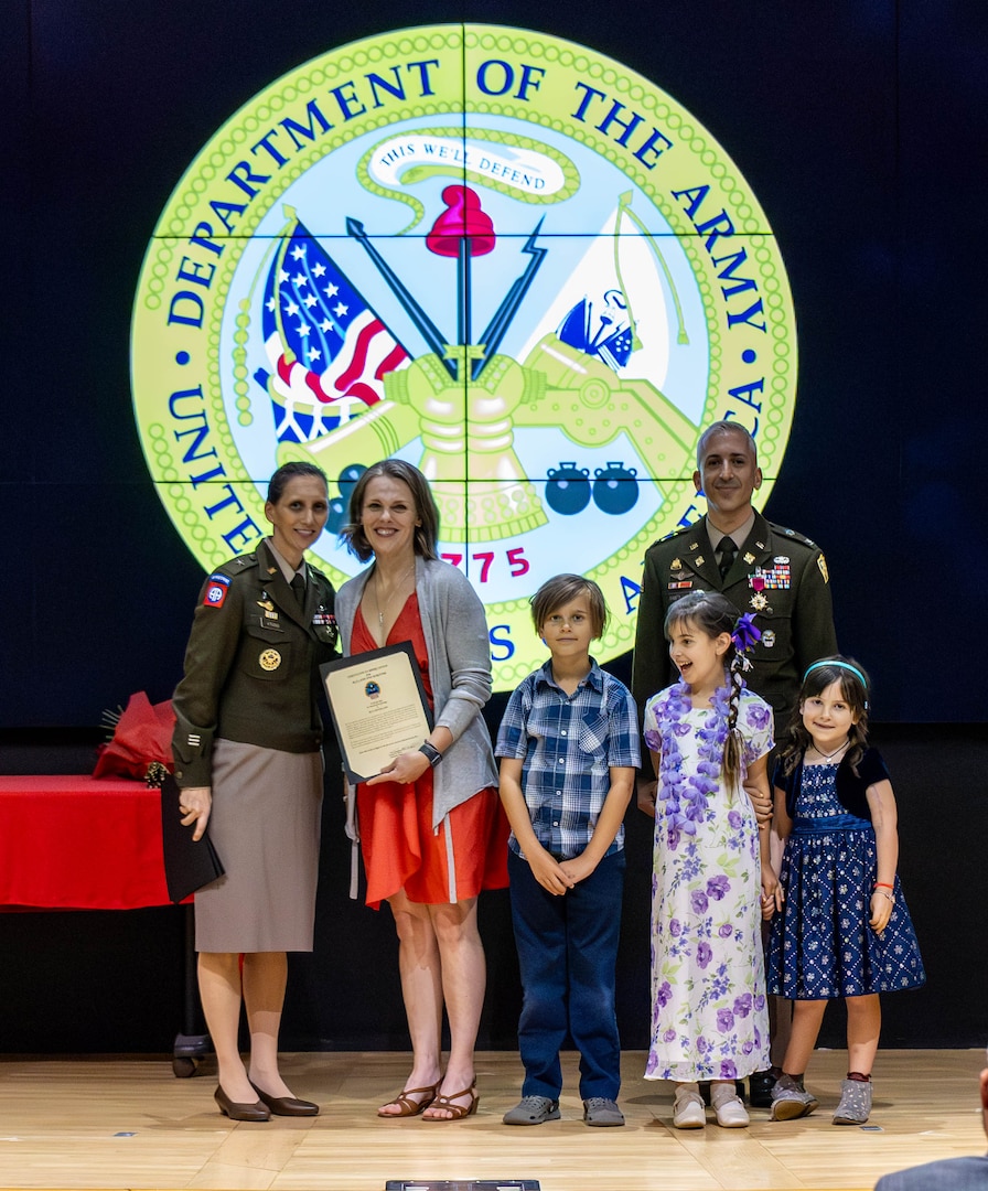 A family stands on stage and are smiling. There are two girls and a boy. A man in uniform and a woman in a pink dress smile as well. Another woman in uniform is smiling next to them.
