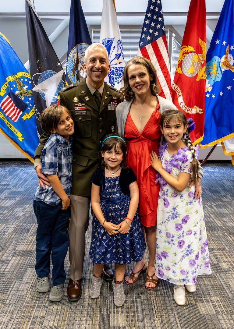 A smiling family stands in front of an array of flags. The man is in uniform.
