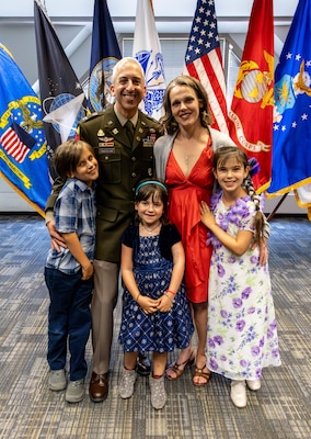 A smiling family stands in front of an array of flags. The man is in uniform.