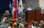 Two people in uniform smile as a girl speaks at a podium behind them.