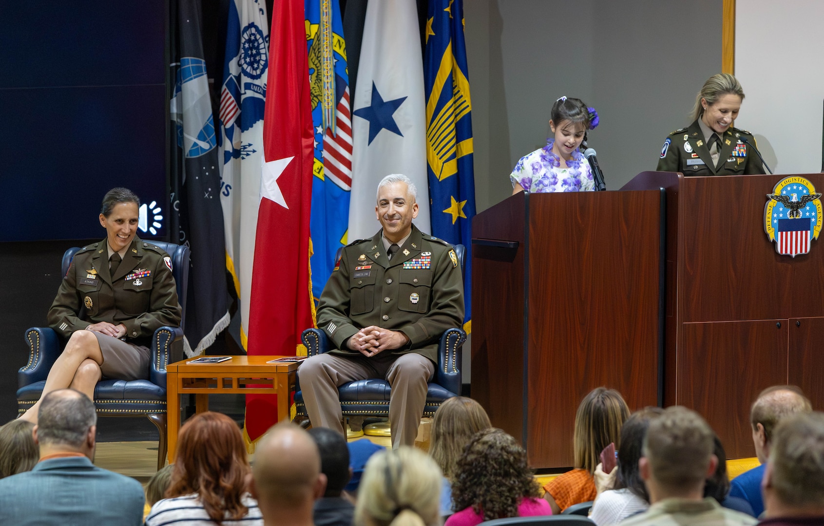 Two people in uniform smile as a girl speaks at a podium behind them.