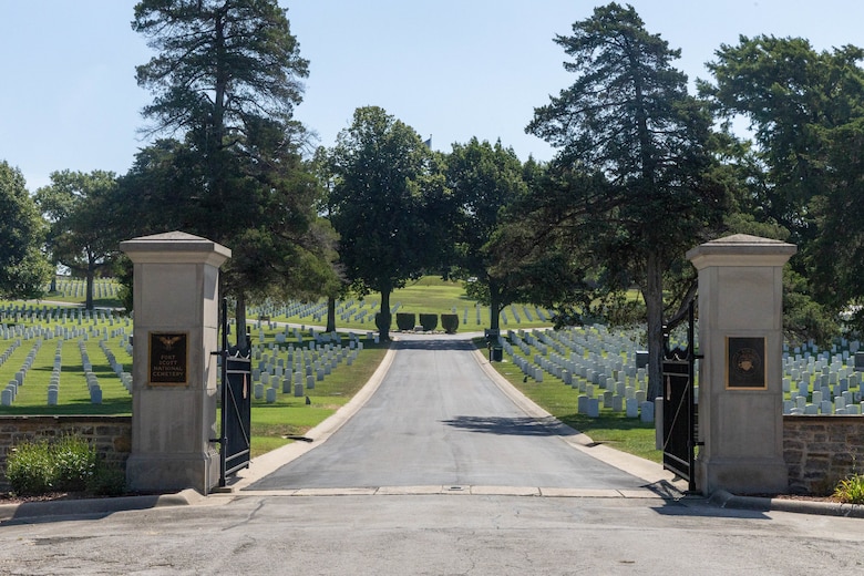 Two grey concrete pillars with black iron gates frame a black road with grass and white headstones on either side and trees in the background.