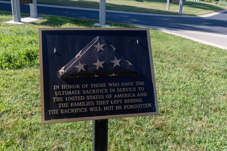 A black sign with a blue folded American flag, gold trim, and gold lettering sits with grass in the background.