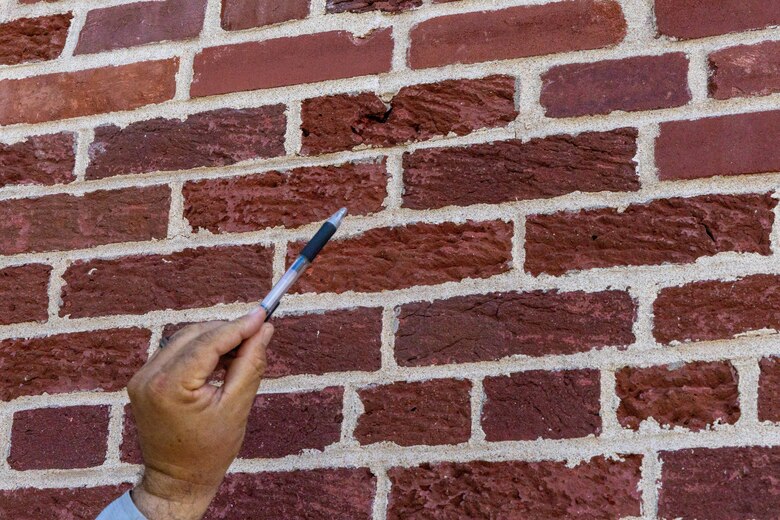 A man's hand holding a pen points to red brick with beige mortar in between the bricks.