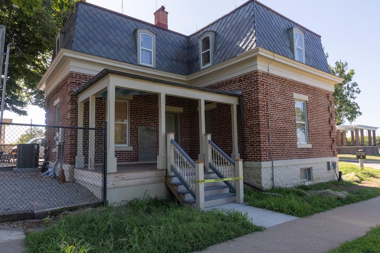 A red brick building with beige trim, a front deck, and a grey slate tile roof with grass in the foreground.