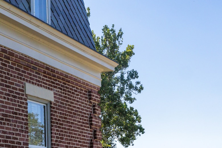 A red brick building with a grey slate roof is on the left with a tree and blue sky on the right.