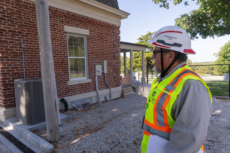 A man in a white hardhat and a yellow safety vest points to a red brick building with grey gravel in the foreground.