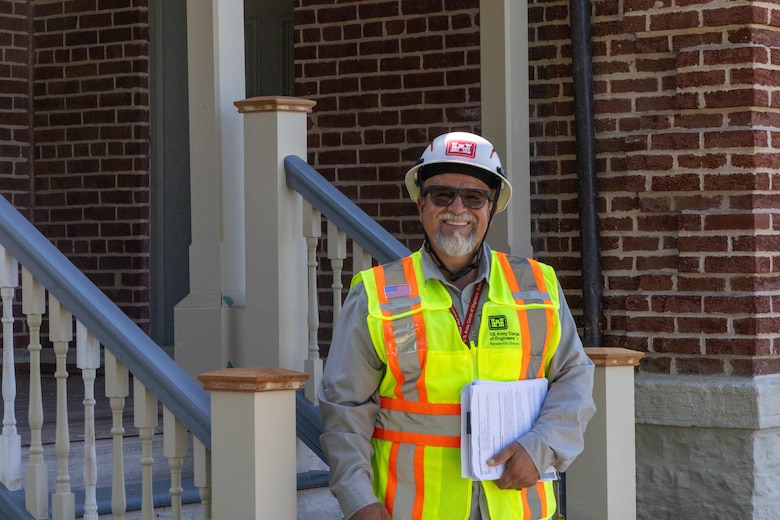 A man in a white hardhat and a yellow safety vest stands and smiles in front of a brick building.