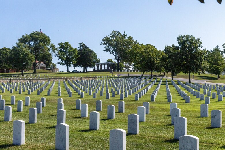 White headstones are lined up in green grass with trees and blue sky in the background.