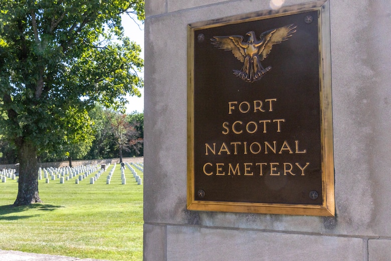 A black sign with gold lettering hangs on a grey concrete post with white headstones, grass and trees in the background.