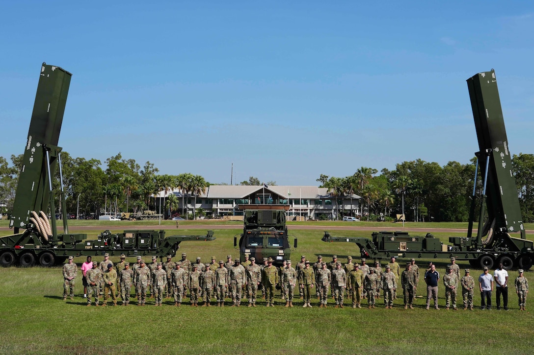U.S. and Australian service members stand together in front of a long-range hypersonic weapon system on a grassy field, with a building and trees in the background.