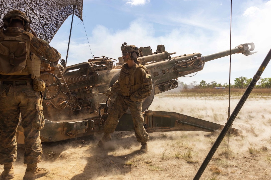 Two Marines fire a large gun in a sandy area with sparse vegetation, with trees in the background on a sunny day.