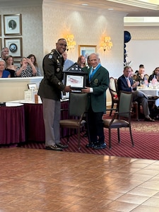 A U.S. Army colonel, in uniform, presents a flag and certificate to an older veteran at a party.