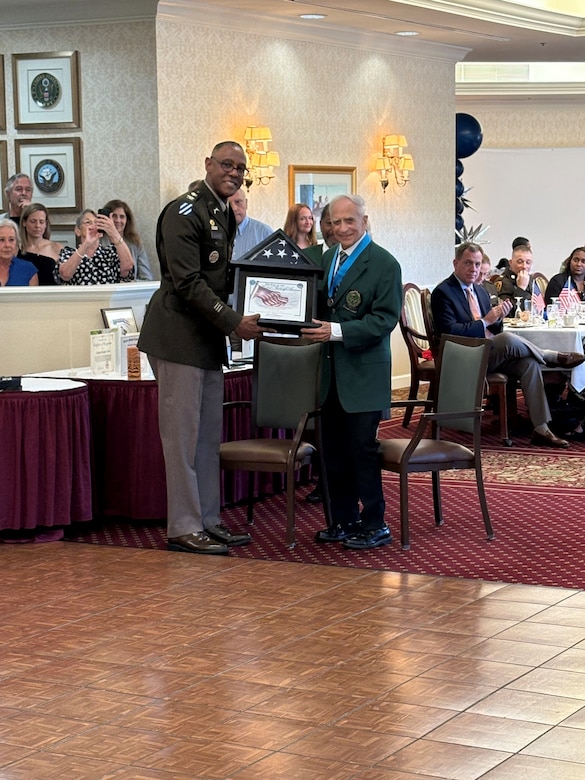 A U.S. Army colonel, in uniform, presents a flag and certificate to an older veteran at a party.