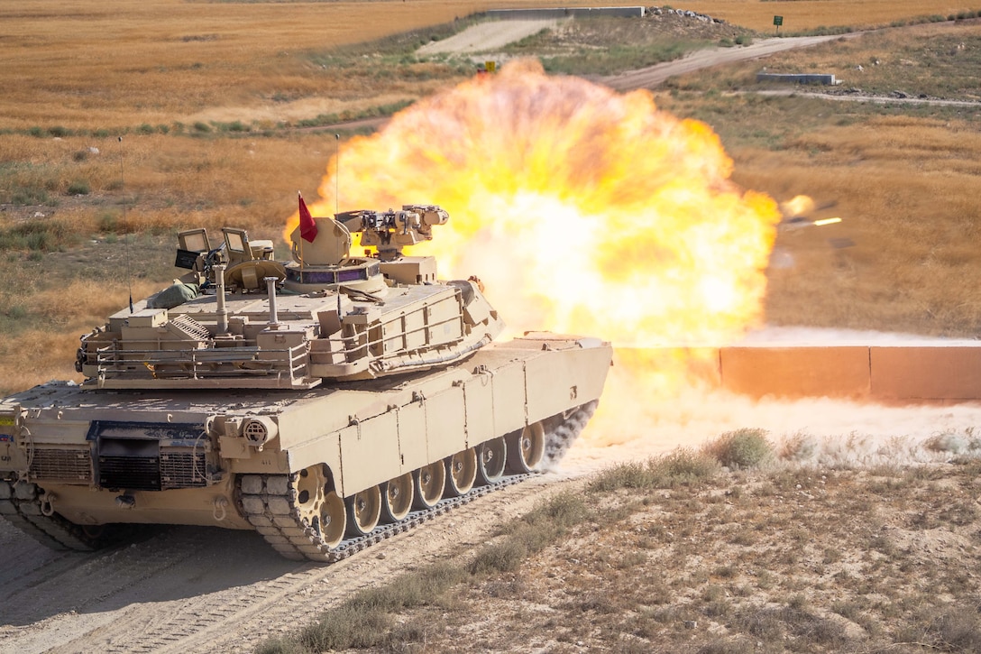 A tank on a dirt road fires toward a field creating an orange fireball.