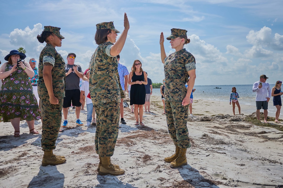 Two Marines raise their right hands on the sand while family members watch in front of the beach.