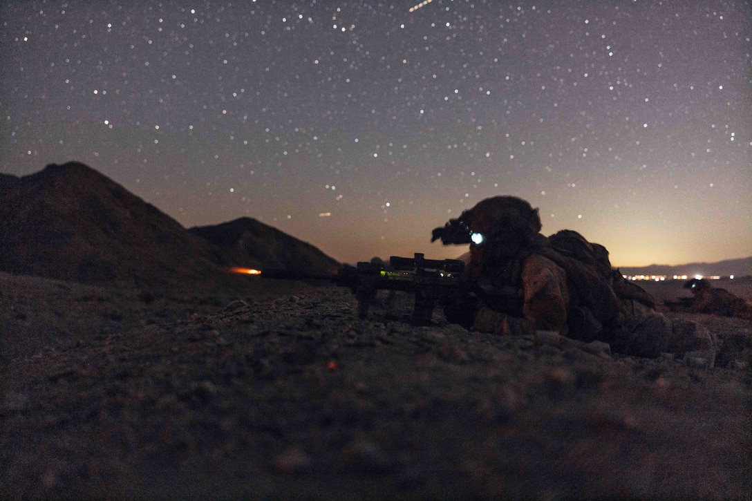 A Marine lying on a rocky surface fires a weapon under a starry sky.