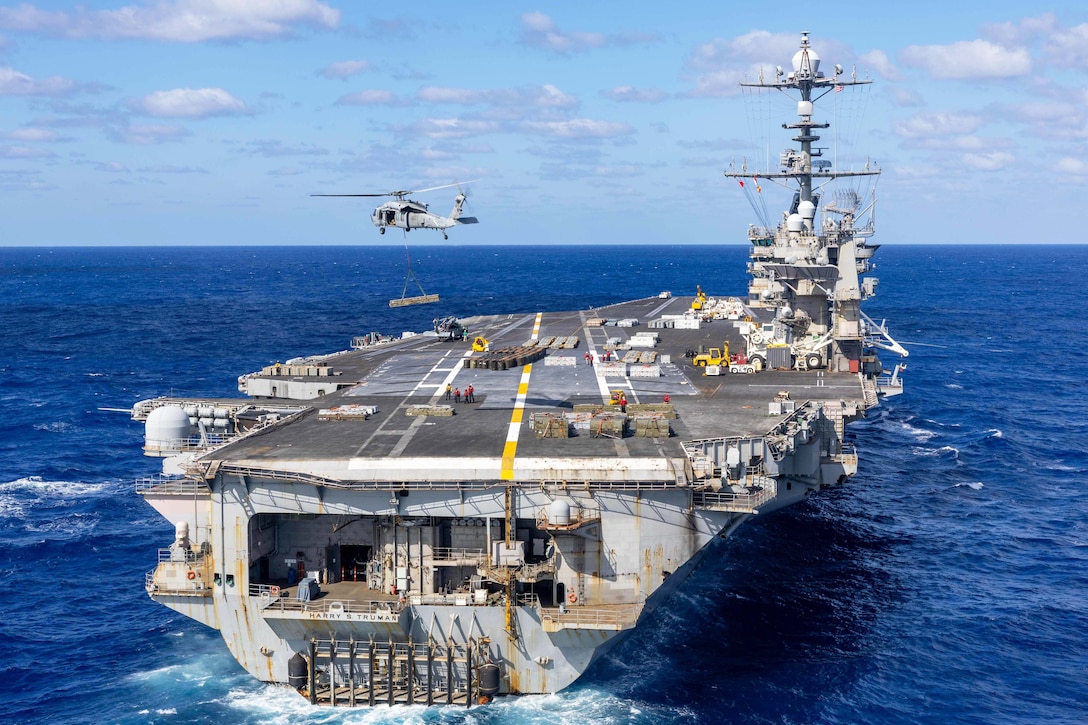 A helicopter carrying ordnance flies over a ship at sea.